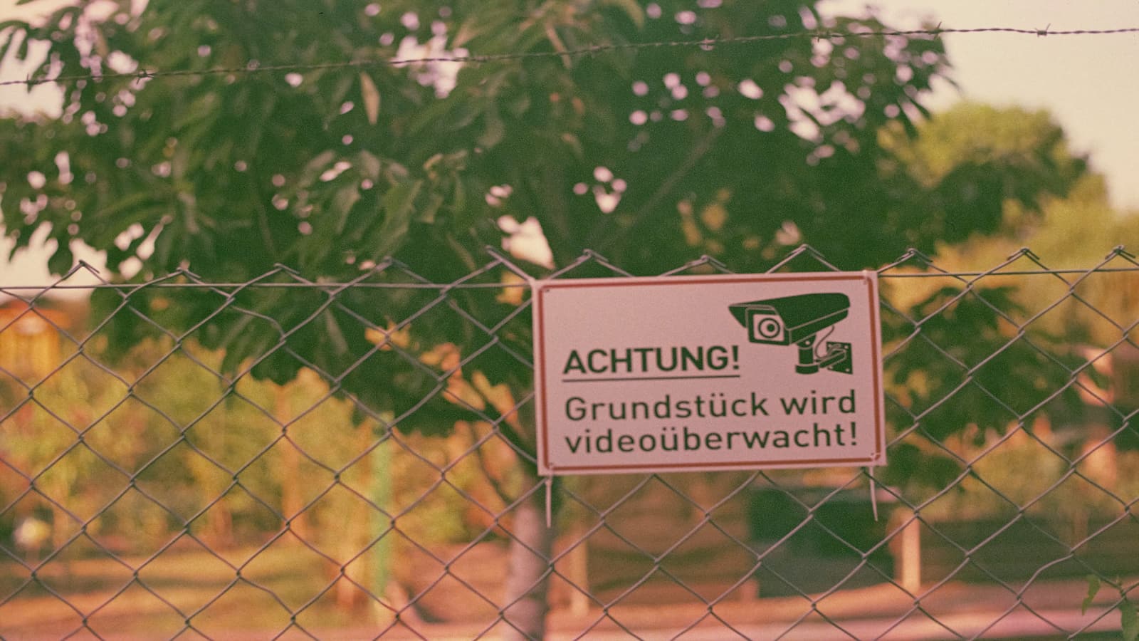 Zaun mit einem Schild, das die Aufschrift 'Achtung! Grundstück wird videoüberwacht!' trägt.